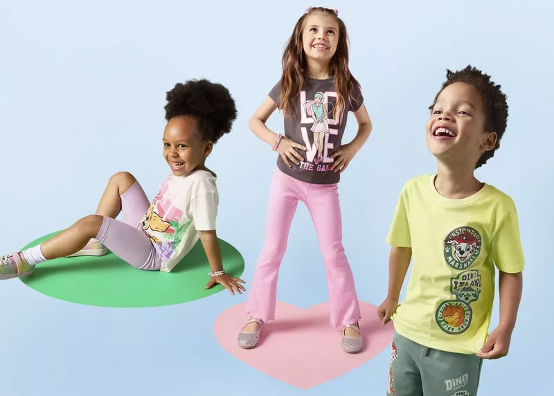 Tre bambini sorridenti indossano abbigliamento casual Pepco con stampe colorate di personaggi dei cartoni animati.